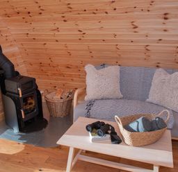 A wooden interior with a small stove and a basket of wood next to a sofa with cushions and a throw and a wooden table with a basket holding rolled towels and binoculars Glamping Pod 5 Shelter Lebberston