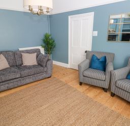 A living room with a grey sofa with beige cushions two striped armchairs with blue cushions and a woven rug at Suffolk House in Narberth