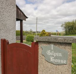 A red wooden gate next to a stone pillar with a sign reading 1 Gilpin House in Levens