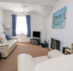 A living room with beige sofas, a small TV on a stand, blue curtains, and a painting on the wall at Bethan Apartment in Trearddur Bay