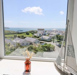 A window view of houses and the sea with a small sailboat model and a jar with dried flowers on a windowsill at Bethan Apartment in Trearddur Bay