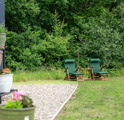 Two green cushioned wooden chairs and a small wooden table on grass near bushes and flower pots on a gravel area at Bedwen in Aberaeron