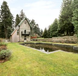 A stone and wood house with a black balcony next to a rectangular reflecting pool surrounded by grass and trees at The Folly in Belford