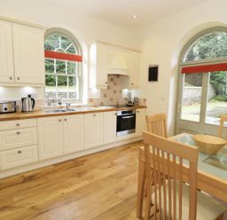 A kitchen with wooden cabinets, countertop appliances, a glass dining table with chairs, and arched windows at The Folly in Belford