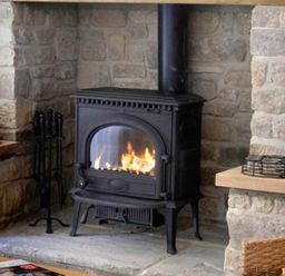 A stone fireplace with a black wood-burning stove fire tools candles and a wicker basket Near Belford