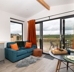 A living area with a blue sofa, orange cushions, a round dining table set for two, and sliding glass doors opening to a balcony with countryside views at Cae Dafadd New Mills near Llanfair Caereinion