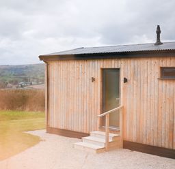 An exterior view of a small wooden cabin with a door and steps overlooking a rural landscape at Cae Bedw in Llanfair Caereinion