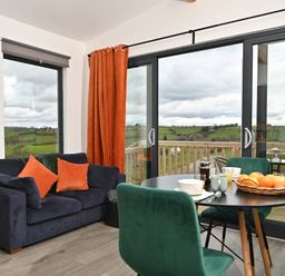 A living area with a dark blue sofa with orange cushions a round dining table with green chairs and glass doors overlooking a green landscape at Cae Bedw in Llanfair Caereinion