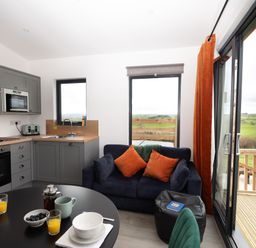 A kitchen and seating area with a round table set with cups and bowls a blue sofa with orange cushions and sliding glass doors opening to a deck with a view at Cae Bedw Llanfair Caereinion