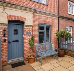 Front door and window of a brick cottage with a blue bench and potted plants at Compass Cottage in Aldeburgh