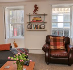 A living room with a brown leather armchair, a grey sofa, a wooden table with pink flowers, and shelves with books and games at The Old Factory House in Berwick-Upon-Tweed