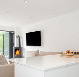 A living room with sofas a wood-burning stove a wall-mounted TV and a kitchen island with coffee and pastries on a tray at 100 Hillside Villas in Looe