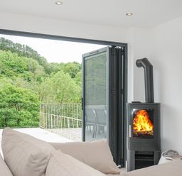 A living room with a beige sofa a black wood stove with fire a wall-mounted TV and an open glass door leading to a balcony with outdoor furniture and a view of green trees at 100 Hillside Villas in Looe