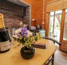 A wooden dining table with a bottle of champagne flowers and glasses in a kitchen with glass doors showing a garden at Snape Castle Garden Lodge in Snape near Bedale