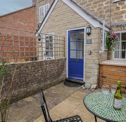 A small outdoor patio area with a blue door in a stone building and a table with a bottle and two glasses at Old Ropemakers Cottage in Bridport