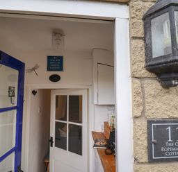 An open blue door leading to a narrow hallway with a white door, a sign on the wall, and a stone exterior wall with a lantern and a plaque reading The Old Ropemakers Cottage at Bridport
