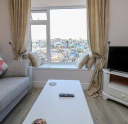 A living room with a grey sofa with fish pillow a white coffee table with remote and coasters TV on white cabinet and large window with cushions and beige curtains at Fishnets in Brixham