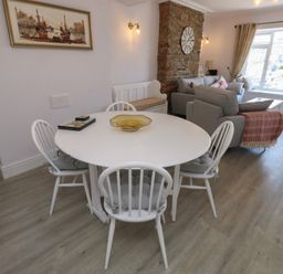 A dining area with a round white table and four white chairs next to a living room with grey sofas and a large window at Fishnets in Brixham