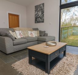 A living room with a grey sofa and patterned cushions a wooden coffee table on a rug and a sliding glass door showing a fenced yard at Silver Birch in Dolton