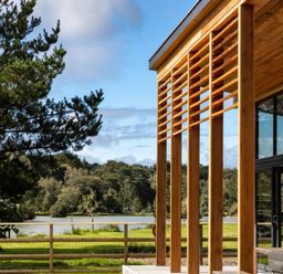 A wooden structure with vertical slats next to glass windows overlooking a lake with trees and grass at Silver Birch in Dolton