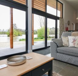 A living room with a gray sofa with animal print cushions and a wooden coffee table with a metal tray and coasters by large glass doors overlooking a lake at Cherry in Dolton