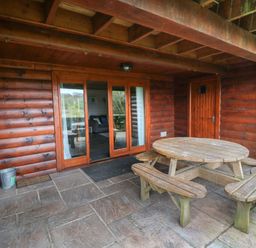A covered patio area with a round wooden picnic table and benches in front of a wooden cabin with glass doors at Aspen Dolton
