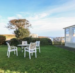 A garden with white plastic chairs and a table next to a caravan and a gate with a view of the coast at Tir Glyn Caravan in Uwch Mynydd near Aberdaron