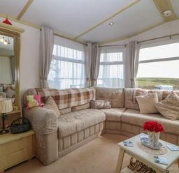 A living room with a beige sectional sofa a small side table with a lamp and radio and a coffee table with a flower vase at Tir Glyn Caravan in Uwch Mynydd near Aberdaron