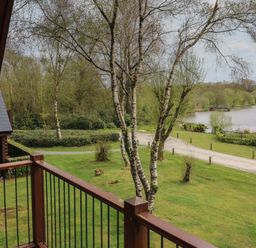 A balcony overlooking trees a grassy area a gravel path and a lake with a wooden building nearby at Cedar in Dolton