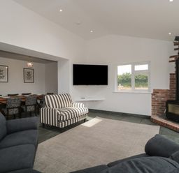 A living room with sofas a striped armchair a wall mounted tv a wood stove and a dining table in the background at Primrose Cottage in Brendon Hill near Roadwater
