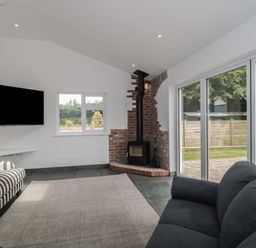 A living room with a TV on the wall a striped armchair a black sofa a brick fireplace and large sliding glass doors at Primrose Cottage in Brendon Hill near Roadwater