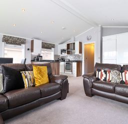 A living room with two brown leather sofas with cushions and an open kitchen area with white cabinets and appliances at Capenwray in Carnforth