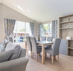 A dining area with a wooden table and six grey upholstered chairs next to windows with patterned curtains and a shelving unit with glassware and a white pot at Coniston in Carnforth