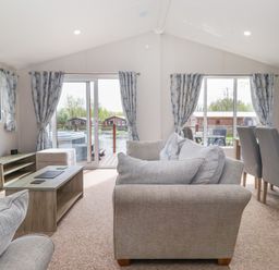 A living room with gray sofas a wooden coffee table and a dining table with chairs near large windows with curtains at Coniston in Carnforth