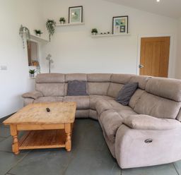 A living room with a beige sectional sofa wooden coffee table and floating shelves with plants and framed pictures at Campion Cottage in Brendon Hill near Roadwater