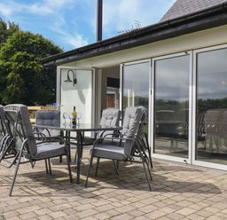 An outdoor patio with a glass table and six cushioned chairs near glass sliding doors at Campion Cottage in Brendon Hill near Roadwater