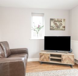 A living room with a brown leather sofa a wooden TV stand with a television a small window with flowers and a painting on the wall at The Brambles in Monmouth