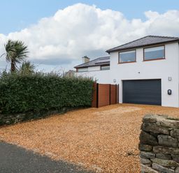 A modern two story house with a black garage door a gravel driveway and a hedge fence at Swyn Llyr in Rhosneigr