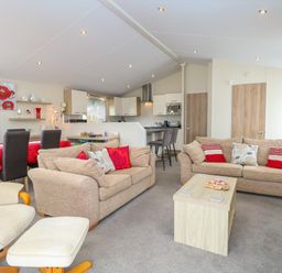 A living room with beige sofas red and patterned cushions a wooden coffee table and dining table with red tablecloth at 21 Norman Drive Lodge in Burgh Castle