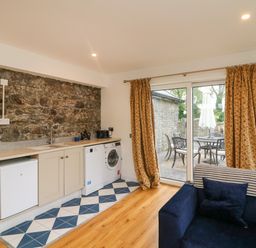 A kitchen area with washing machine fridge and sink next to sliding glass doors with yellow floral curtains and a blue sofa with pillows at The Farm Cottage in Athy County Kildare