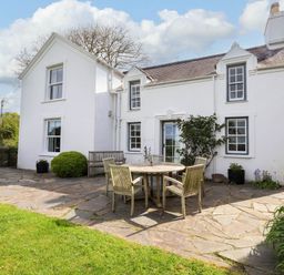 A white house with windows and a chimney with a stone patio with a round table and chairs and green lawn at Bryn Tirion in Morfa Nefyn