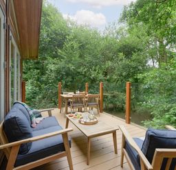 An outdoor wooden deck with cushioned chairs a table with drinks and food surrounded by trees at 10 Stonerush Valley Lanreath near Looe