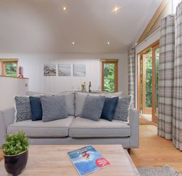 A living room with a grey sofa cushions a coffee table with a book a plant large windows with plaid curtains at 10 Stonerush Valley in Lanreath near Looe