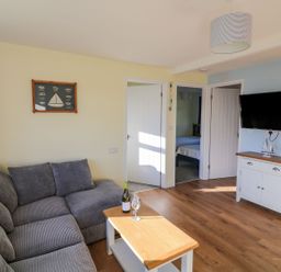 A living room with a gray sofa a wooden coffee table a wall-mounted television and an open door showing a bedroom at The Sail Loft in Norton Park near Dartmouth