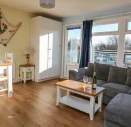 A living room with a gray sofa a wooden coffee table with wine bottle and glasses a dining table with chairs a wardrobe and nautical decor at The Sail Loft in Norton Park near Dartmouth