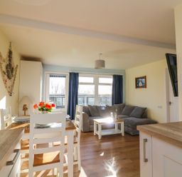 A dining area with a wooden table and white chairs next to a living room with a grey sofa and coffee table at The Sail Loft in Norton Park near Dartmouth