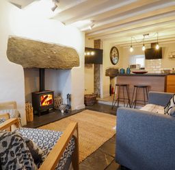 A living room with a wood burning stove two armchairs a gray sofa and a kitchen area with stools and hanging lights at Ty Dyffryn in Llan Ffestiniog