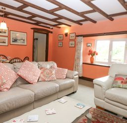 A living room with a beige sofa and armchair with patterned cushions a coffee table and framed pictures on orange walls at Loanview in Cornhill-On-Tweed