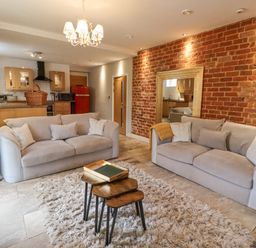 A living room with two beige sofas a wooden nesting table and a chandelier with an open kitchen and exposed brick wall at The Fullerton in Belsay