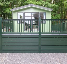 An outdoor view of a light green cabin with a dark green fenced deck surrounded by trees at Eltons Escape in Pony Meadow 3 White Cross Bay near Windermere